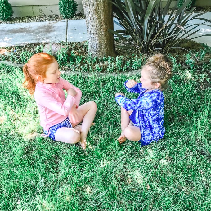 Two girls sitting on grass in their swimsuits, playing pat-a-cake.