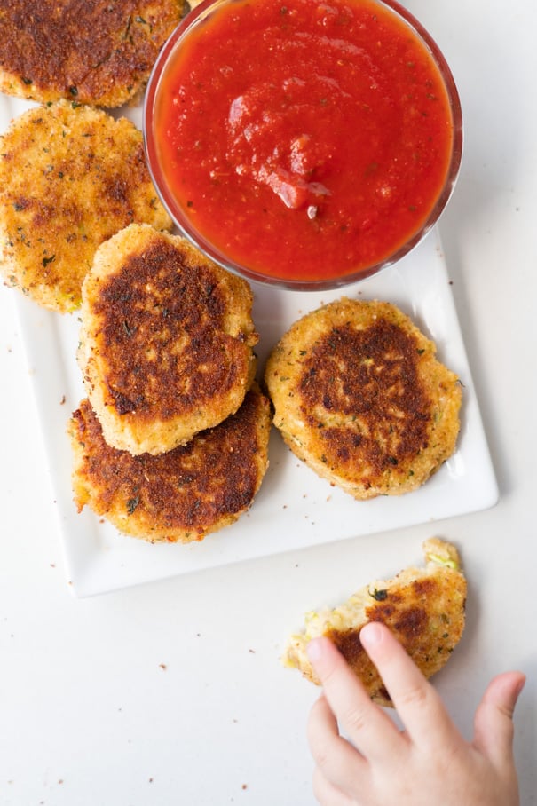 A white plate full of zucchini tots and a small hand reaching to grab one.