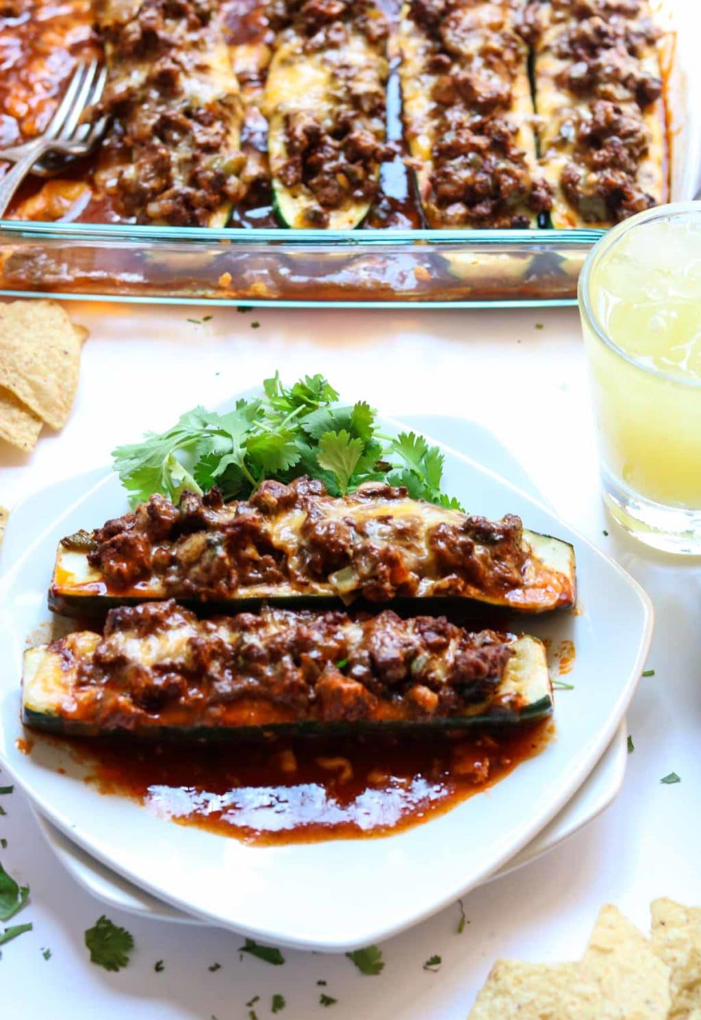 Two enchilada zucchini boats on a white plate in front of a glass baking dish.