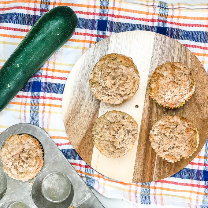 Four zucchini muffins with streusel topping on a round piece of wood over colorful kitchen towel.
