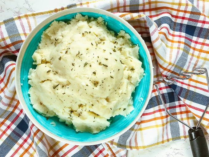 A teal bowl filled with instant pot garlic mashed potatoes next to a potato masher.