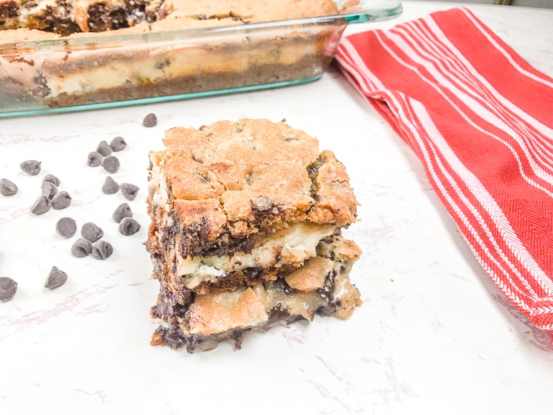 Chocolate chip cookie cheesecake bars stacked up in front of pan of bars.
