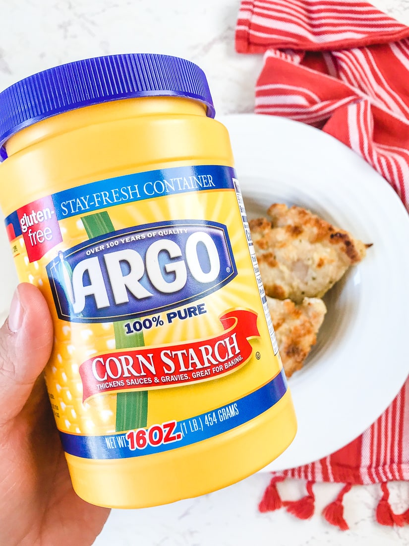 A hand holding up a container of Argo Corn Starch over a bowl of crispy oven baked chicken thighs.