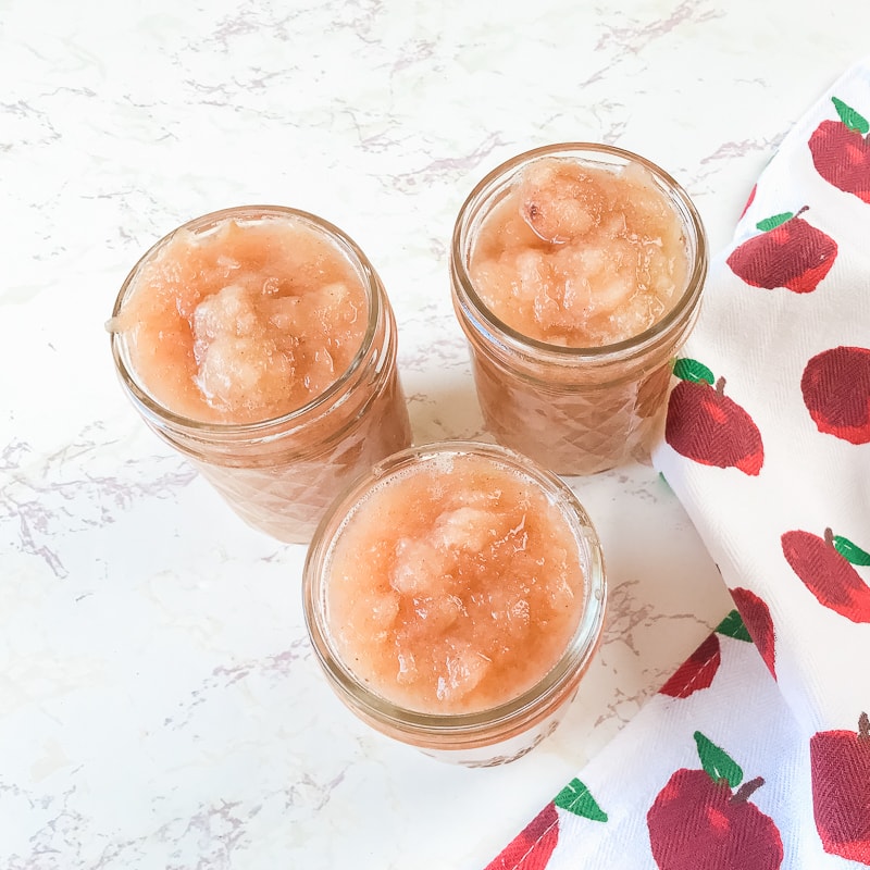 Three jars of instant pot applesauce next to an apple printed towel.