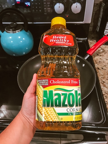 A hand holding a bottle of Mazola Corn Oil in front of a stove.