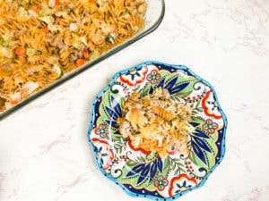 A casserole dish filled with turkey noodle casserole next to a colorful plate with a serving of turkey and noodle casserole.