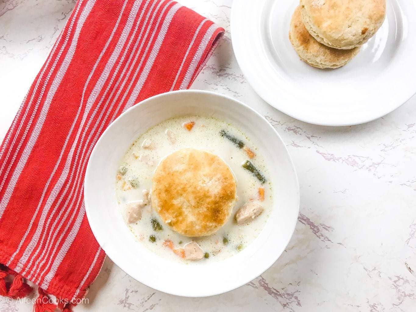 A white bowl filled with instant pot chicken pot pie soup and topped with a biscuit next to a red striped towel.