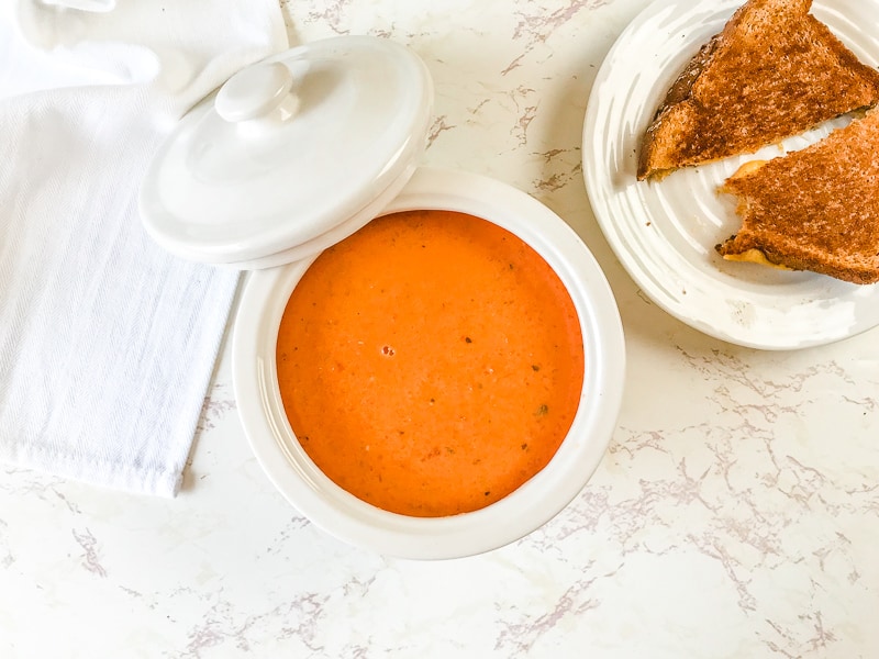 A white bowl of tomato basil soup next to a grilled cheese sandwich.
