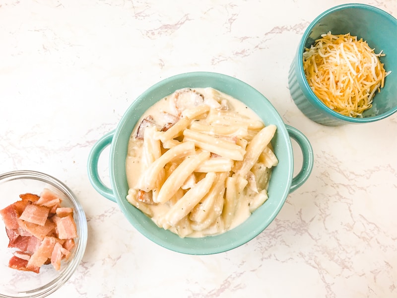 A bowl of cheesy pasta next to small bowls of shredded cheese and bacon.