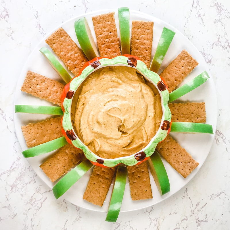 A serving platter filled with sliced green apples, graham crackers, and a bowl of pumpkin pie dip in the centenaries.