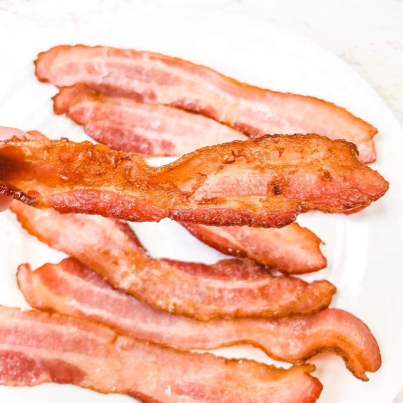 A slice of extra crispy air fryer bacon being held in front of a plate of bacon.