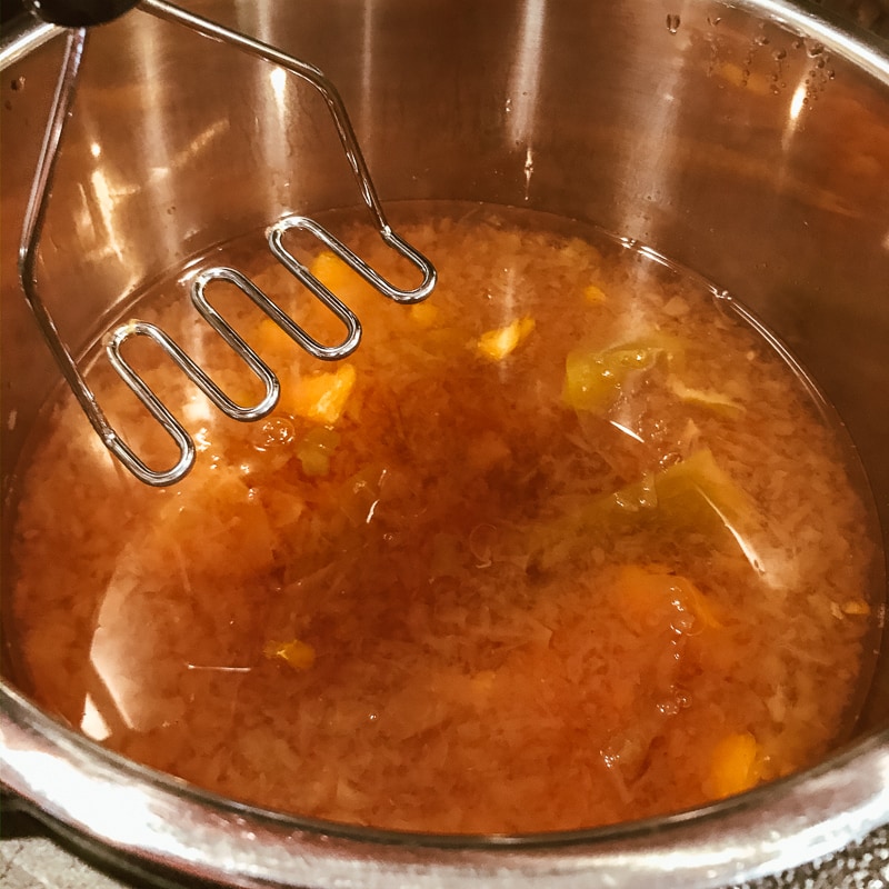 A potato masher mashing up the fruit inside the pot of homemade apple cider.