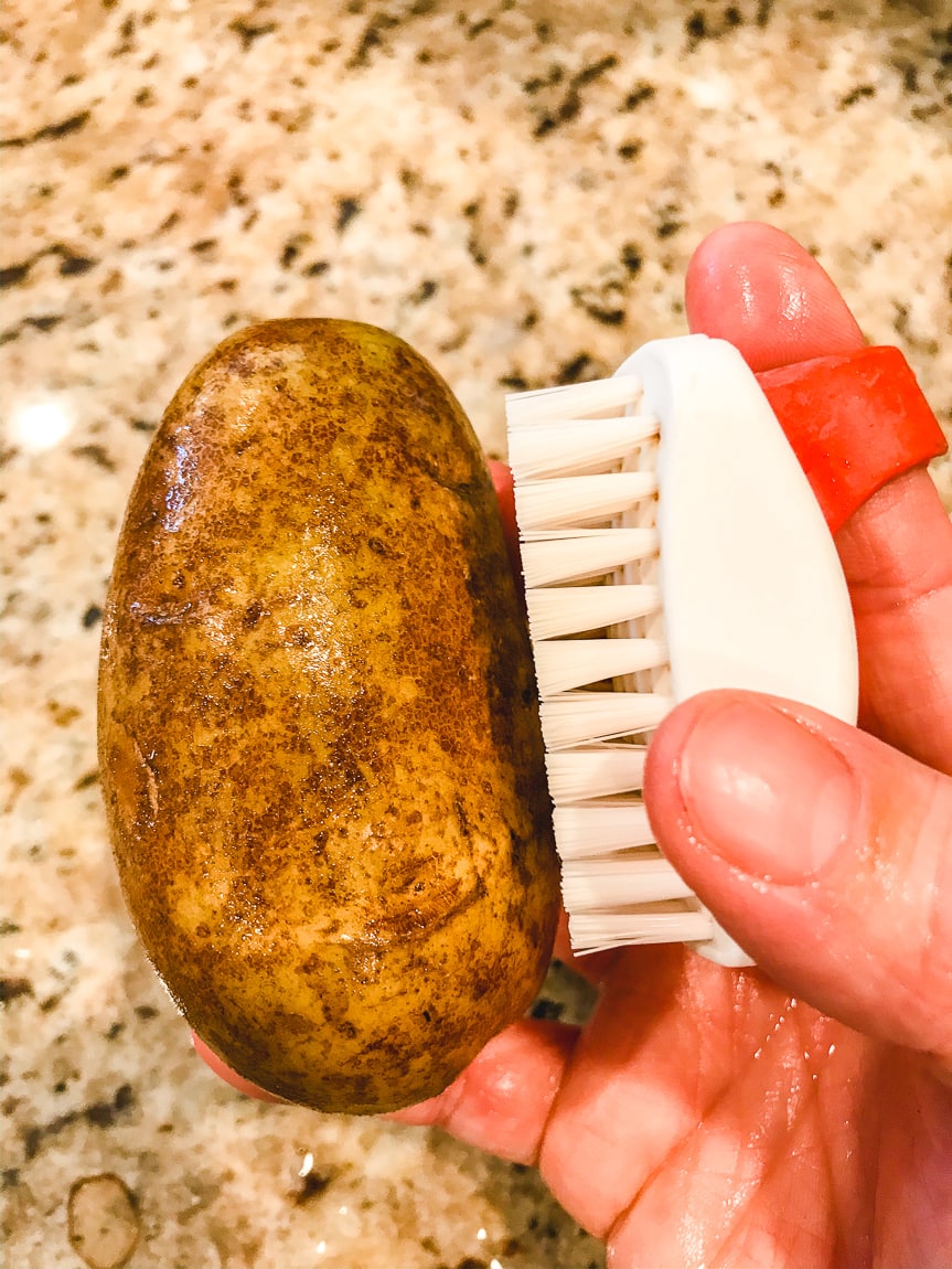 A hand scrubbing a russet potato with a red and white potato scrubber.