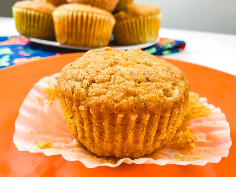 A pumpkin cornbread muffin on an orange plate with the liner peeled off to show the lines of the pumpkin muffin underneath.