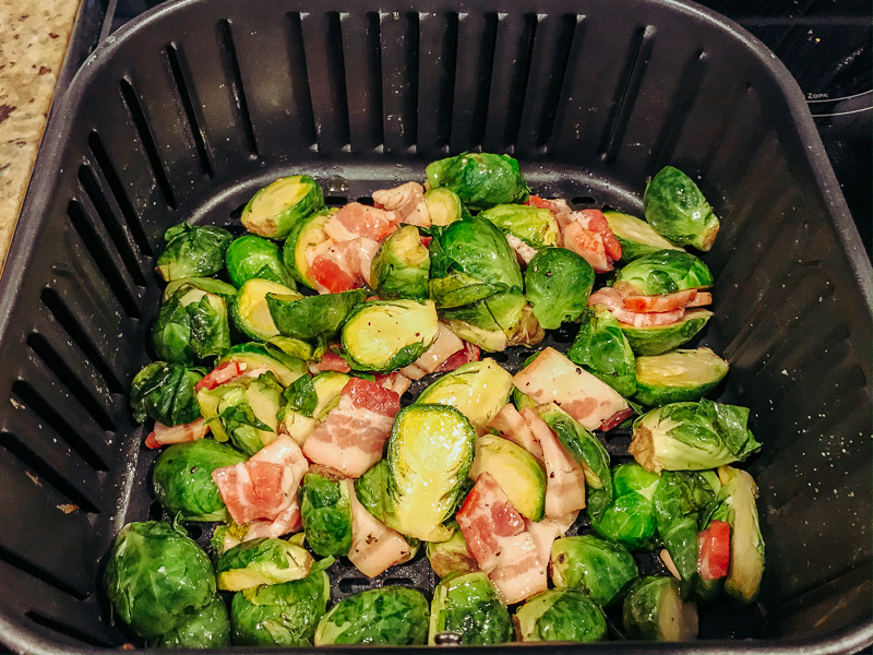 Raw brussels sprouts and bacon inside basket of air fryer.