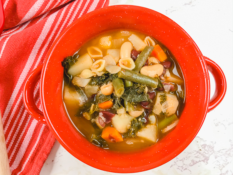 Overhead shot of minestrone soup in red bowl next to red striped towel.