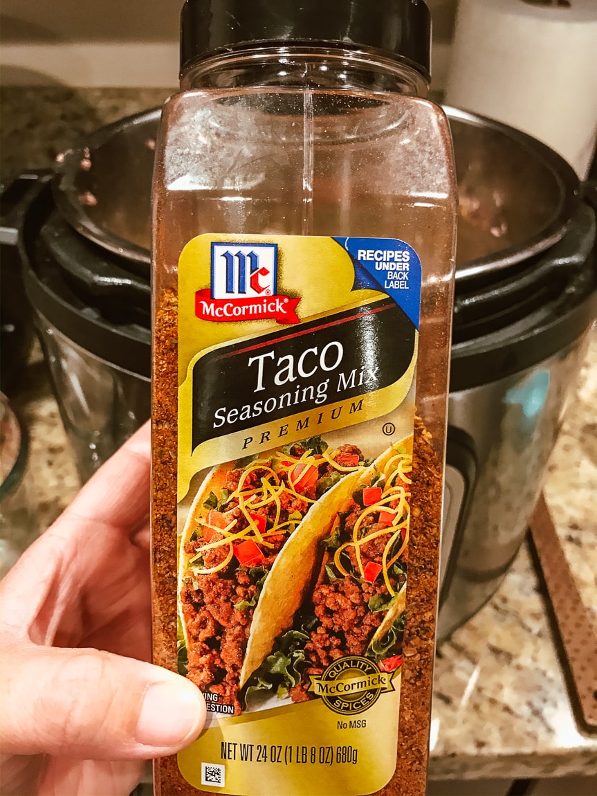 A hand holding up a big bottle of McCormick taco seasoning in front of an Instant Pot.