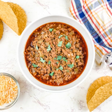 A white bowl filled with instant pot taco meat and surrounded by crispy taco shells and a small dish of shredded cheese.
