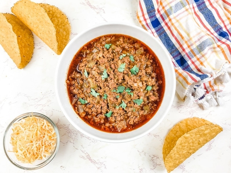 A white bowl filled with instant pot taco meat and surrounded by crispy taco shells and a small dish of shredded cheese.