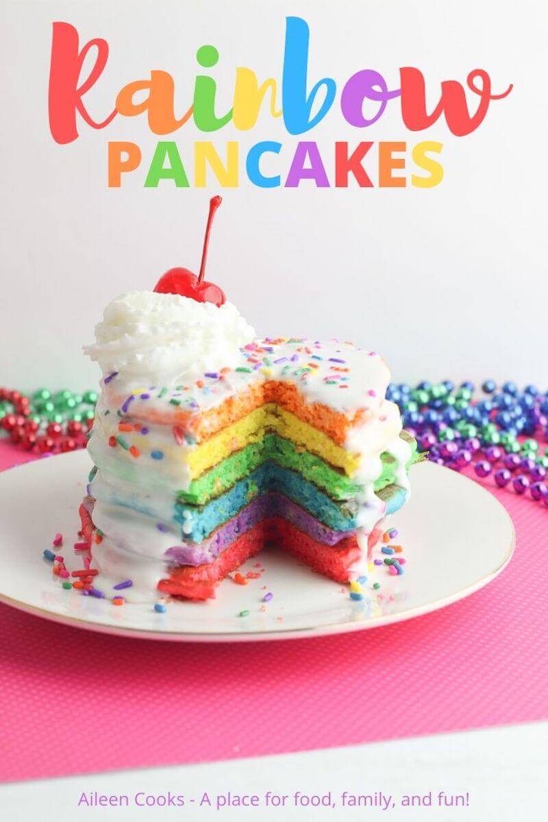 A tall stack of rainbow colored pancakes with whipped cream and cherry on top.