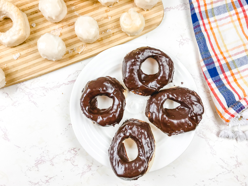 A white plate of chocolate glazed donuts next to a cutting board filled with glazed donuts and donut holes.