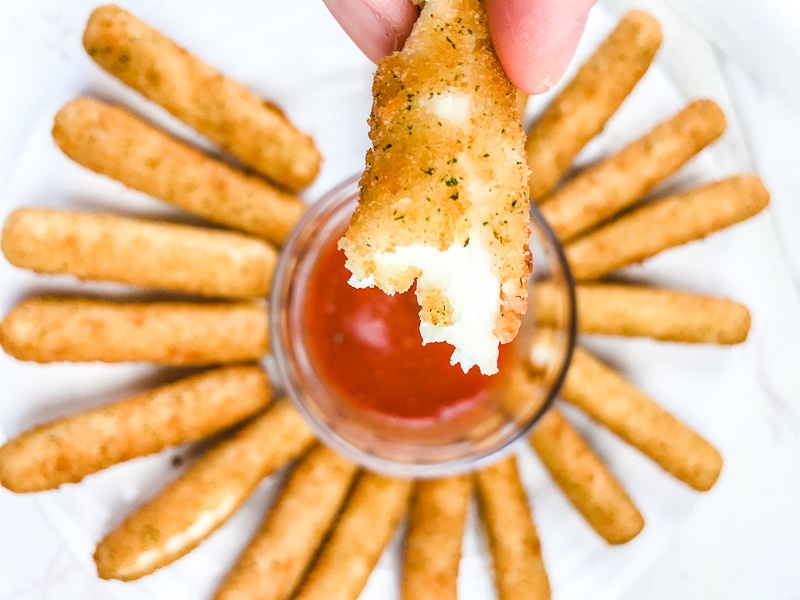 A mozzarella stick with a bite out of it above a plate of sticks and red marinara sauce.