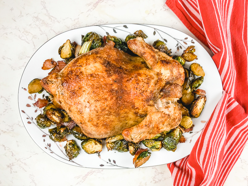 Whole chicken on white platter surrounded by roasted Brussels sprouts.