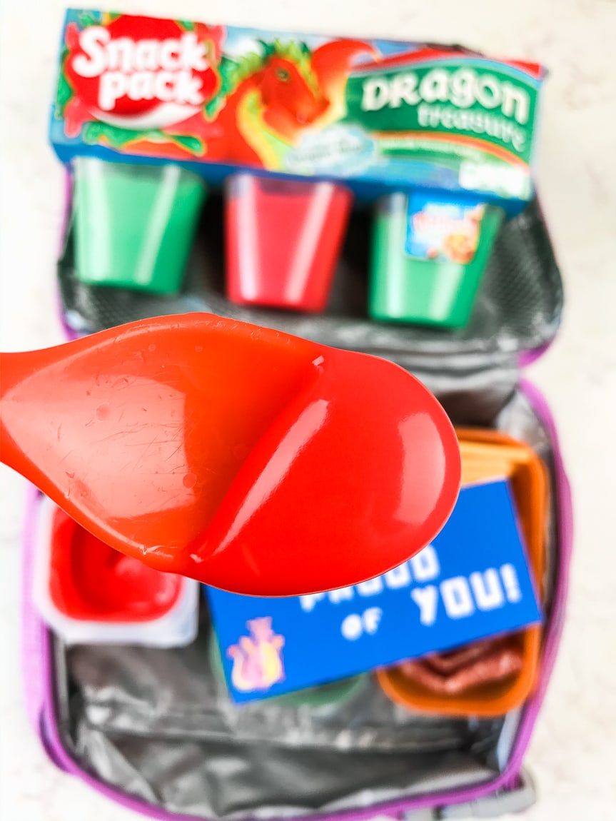 Spoonful of red snack pack above lunchbox.