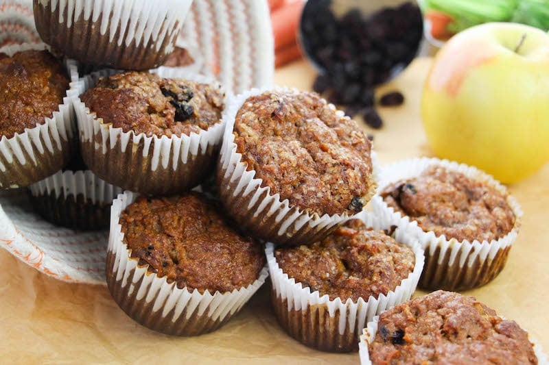 A pile of morning glory muffins spilling out of a bowl with an apple and raisons in the background.