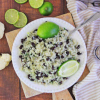 White dish of cauliflower rice next to cauliflower florets, limes, and striped towels.