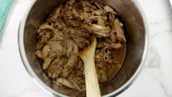 Pork being cooked with additional liquid and spices and stirred with wooden spoon.