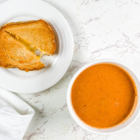 A bowl of tomato soup next to a grilled cheese cut in half.