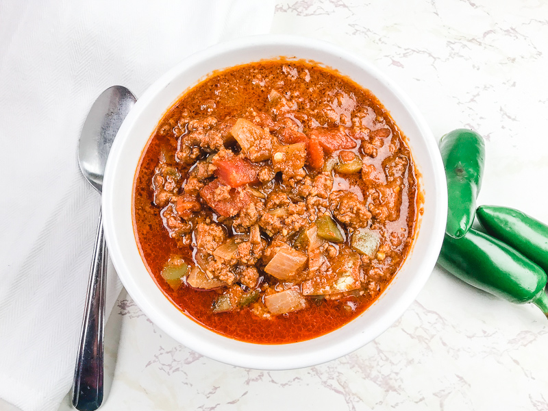 A white bowl of no bean chili next to three jalapeños.