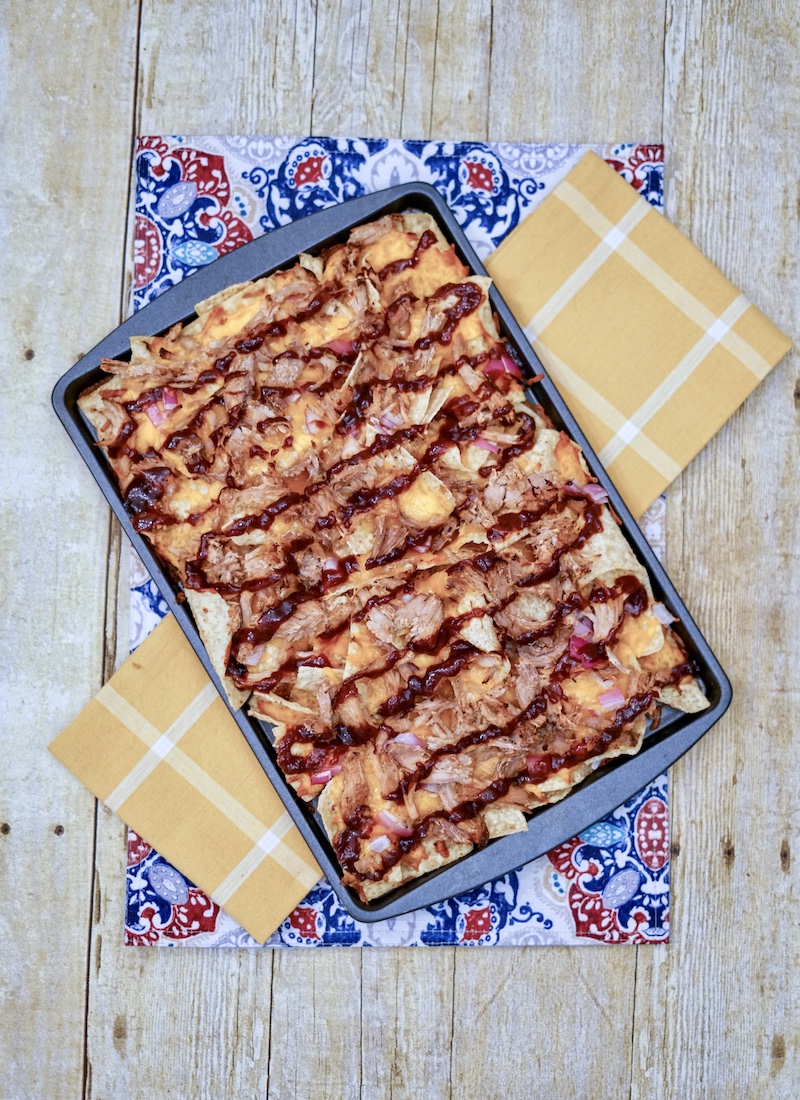 A cookie sheet filled with pulled pork nachos on top of blue and yellow dish cloths.