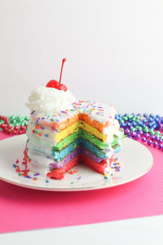 A tall stack of colorful rainbow pancakes with frosting and sprinkles.