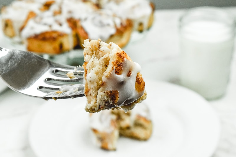 A bite of cinnamon rolls on a fork in front of a plate of cinnamon rolls and glass of milk.