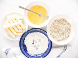 Three bowls with flour, breadcrumbs, and eggs, next to a plate of cheese sticks.