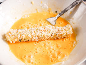 A breaded cheese stick being dipped into beaten eggs again.