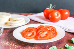 Slices of tomatoes on a white plate with slices of mozzeralla on a white plate behind the plate of tomatoes.