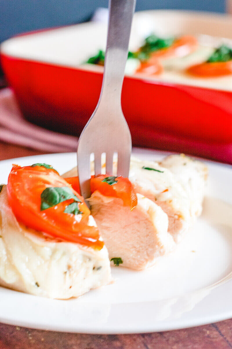 A fork sticking out of a slice of chicken breast topped with mozzarella, tomato, and basil.