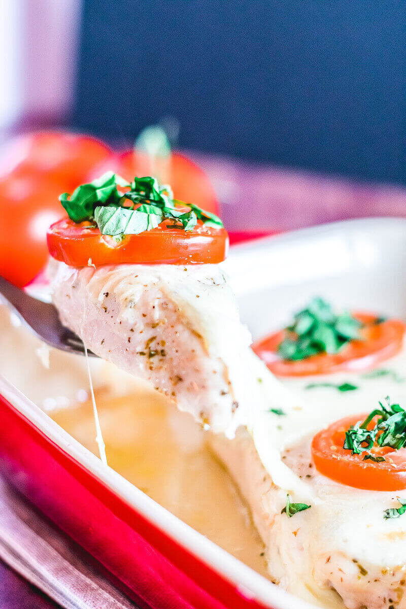 A piece of baked chicken caprese being lifted out of a red baking dish.