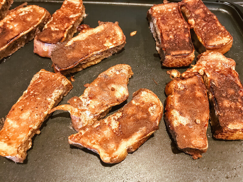 French toast sticks on a griddle, having already been flipped and cooked on one side.