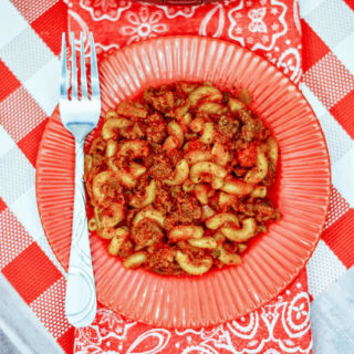 A red plate of goulash on top of a checkered tablecloth.