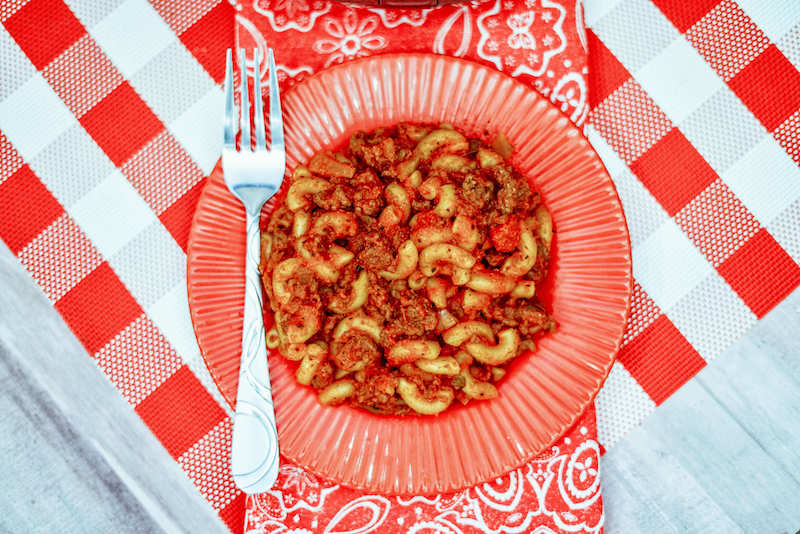 A red plate of goulash on top of a checkered tablecloth.