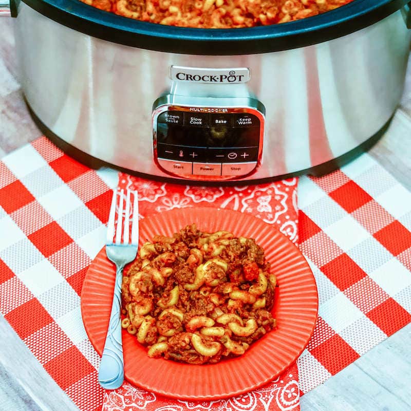 A red plate filled with goulash with a fork next to it and on top of a red checkered placemat.