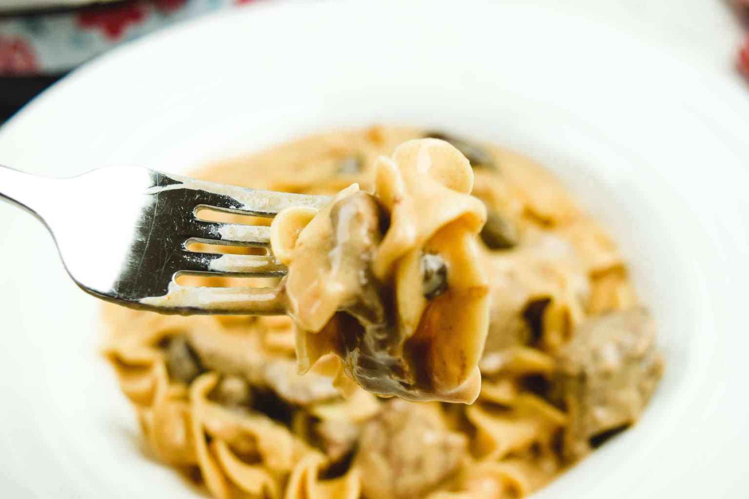 A bite of stroganoff on a fork in front of a bowl of stroganoff