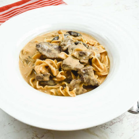 Side angle view of beef stroganoff in a wide rimmed bowl next to a red striped dish towel.