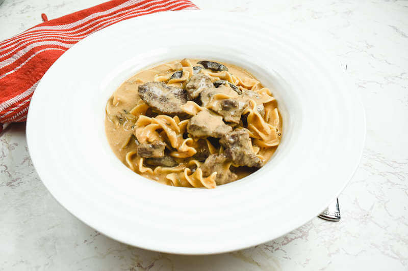 Side angle view of beef stroganoff in a wide rimmed bowl next to a red striped dish towel.