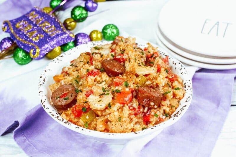 A white bowl filled with jambalaya next to Mardi Gras decorations in purple, gold, and green.