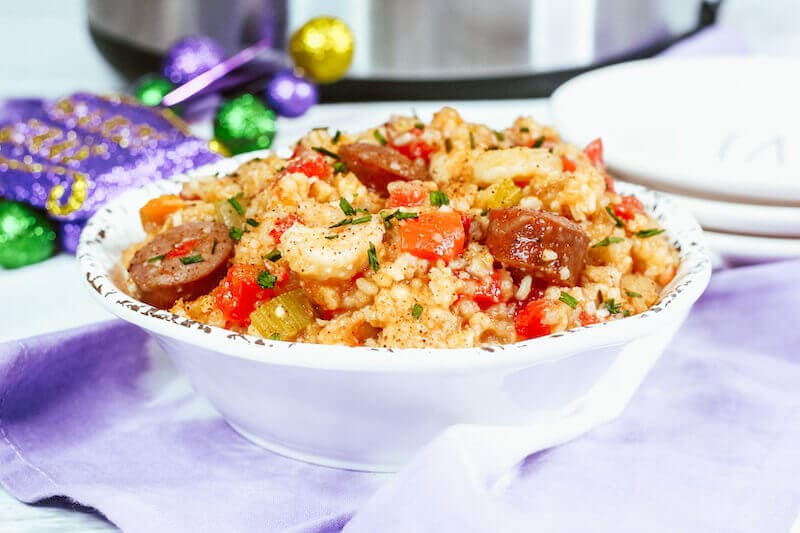 A white bowl of jambalaya next to Mardi Gras decorations in purple and an instant pot.